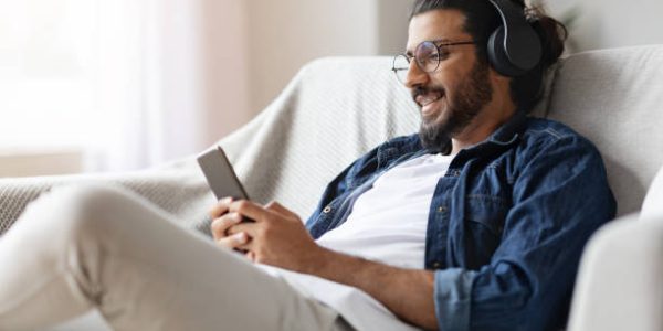 Home Leisure. Young Western Guy In Wireless Headphones Relaxing With Smartphone On Couch, Listening Music And Messaging With Friends, Enjoying Domestic Pastime, Resting On Comfortable Sofa, Free Space